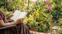 Senior woman with brown skin reading an open book in a lush garden surrounded by blooming flowers and green foliage during daytime, suitable for lifestyle and wellness content.