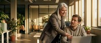 Senior woman with gray hair and light skin tone, wearing a brown blazer, leaning over a seated middle-aged man with light skin tone in a beige shirt, both smiling while working together at a laptop in a modern office with plants and natural light.