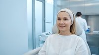Middle-aged Caucasian female patient in white medical gown and headband smiling in modern hospital room with healthcare professional in background.
