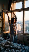 Woman with long dark hair, appearing to be in her twenties to thirties of mixed ethnicity, stretching arms above head by large bedroom window at sunrise, wearing casual brown shirt and blue pants, overlooking cityscape. Morning routine and wellness concept.