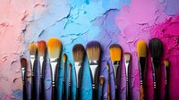 Professional makeup brushes arranged in row on colorful textured background with pink, blue and purple paint strokes creating artistic composition.