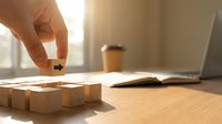 Hand placing wooden cube block completing puzzle on desk with laptop and coffee cup in warm sunlight creating business strategy concept.