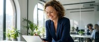 Professional middle-aged Caucasian businesswoman with curly hair smiling while working on laptop in modern office environment with colleagues in background.