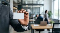Businessman in gray suit holding blank white business card in modern office environment with colleagues working in background.