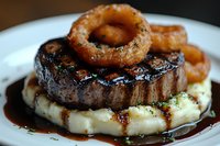 Grilled beef steak topped with crispy onion rings served on creamy mashed potatoes with rich brown gravy sauce on white plate.