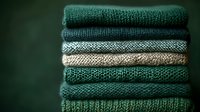 Stack of cozy knitted sweaters in green and beige tones folded neatly, showcasing various knit patterns and textures for autumn winter fashion wardrobe.