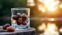 Fresh dates soaking in clear water glass on wooden surface against golden sunset background, natural food photography with bokeh effect.