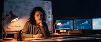 Young African American woman software developer working late night coding session at computer workstation with multiple monitors and planning boards.