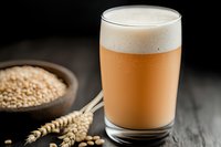 Fresh craft beer glass with creamy foam head next to wheat grains and ears on dark wooden table, selective focus on beverage against black background.