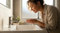 Woman with dark hair and olive complexion washing hands at bathroom sink near window with natural light and potted plant, promoting hygiene and wellness routines.