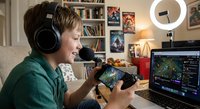 Young male gamer streaming video games with headphones and controller in cozy home setup with ring light and laptop for content creation.