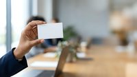 Young professional man with medium brown skin tone displaying blank white business card in modern office environment with wooden desk and blurred background.