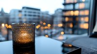 Lit candle in textured glass holder on table with blurred city skyline and illuminated buildings at dusk, suitable for home decor and lifestyle content.