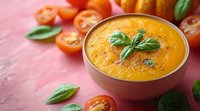 Fresh homemade tomato soup garnished with basil leaves and black pepper, served in ceramic bowl on pink background with cherry tomatoes and herbs.