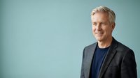Middle-aged man with gray hair, Caucasian ethnicity, wearing dark blazer and navy shirt against turquoise background. Corporate professional portrait for business, leadership, and executive use.
