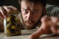 Bearded man with intense expression examining jar of homemade pickles, close-up portrait showing curiosity and concentration during food preservation process.