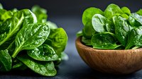 Fresh raw spinach leaves with water drops in wooden bowl and scattered on dark background, macro food photography for healthy eating concept.