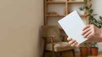 Adult holding blank white tablet or clipboard with both hands against neutral beige wall with wooden shelving and potted plants in background, suitable for mockups and branding presentations.