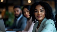 Young African American businesswoman with curly hair and mint blazer smiling confidently during office meeting with diverse colleagues working on laptops in modern workspace.