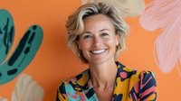 Cheerful middle-aged blonde woman with bright smile wearing colorful floral shirt against orange wall with tropical plant decorations, expressing joy and confidence.