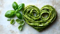 Fresh pasta shaped as heart with green basil pesto sauce and aromatic herbs on light marble background, romantic Italian cuisine concept.