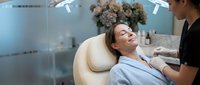 Young woman relaxing during professional facial treatment at modern spa salon with beautician performing skincare therapy in wellness center.