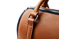 Close-up detail of brown leather handbag showing textured grain, contrast stitching, and metal hardware against white background, highlighting quality craftsmanship.