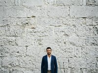 White brick wall background with young man wearing navy blazer and white shirt standing centered for corporate portrait or professional headshot use.