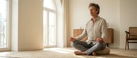 Middle-aged Caucasian man practicing mindfulness meditation in bright modern living room with natural sunlight streaming through large windows.