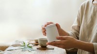 Unrecognizable person holding blank white cup on wooden table with scattered items, suitable for beverage branding and lifestyle marketing.