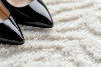 Black patent leather stiletto shoes on white fluffy carpet, creating contrast between luxury fashion and soft texture. Close-up view captures reflection and detail.