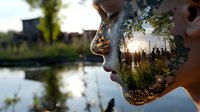 Double exposure profile of young woman combined with sunset nature scene showing silhouettes of people and trees reflected in calm lake waters.