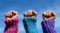 Raised fists wearing colorful knitted wool wristbands against blue sky background symbolizing unity, solidarity, protest and women empowerment movement.