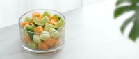 Mixed diced vegetables in glass bowl on white surface with plant leaf, ready for cooking or meal preparation.