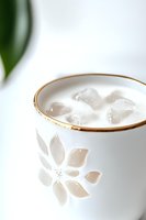 White ceramic cup with floral pattern and gold rim containing ice cubes, photographed in soft light against minimal background. Elegant drinkware for beverage styling.