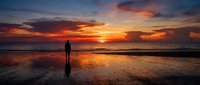 Silhouetted figure standing on wet beach at dramatic sunset with golden and orange sky reflecting on water, contemplative mood.