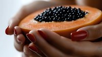 Fresh ripe papaya half filled with black caviar held in female hands with red manicure against blurred background, luxury gourmet food concept.