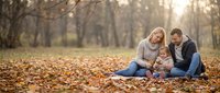 Family of three sitting together on autumn leaves in park, parents with young child enjoying outdoor time during fall season.
