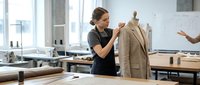 Young female fashion designer adjusting beige coat on mannequin in modern tailoring workshop studio with natural lighting and professional equipment.