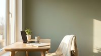 Minimalist home office workspace with wooden desk, laptop, plant and chair against sage green wall with natural sunlight streaming through window.