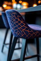 Modern bar stool with geometric orange pattern on dark blue velvet upholstery, bokeh lights in background creating luxurious restaurant atmosphere.