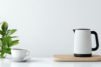 Modern white electric kettle on wooden board with coffee cup and green plant against light wall, minimalist kitchen counter setup for morning routine.