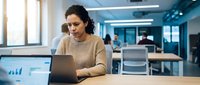 Woman of South Asian descent, approximately 30s, wearing a beige sweater, focused on laptop at desk in modern office with blue accents and colleagues working in background.