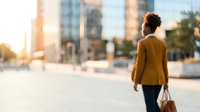Woman with curly hair wearing mustard yellow jacket walking through urban downtown street with modern skyscrapers, suitable for business, lifestyle, and city living concepts.