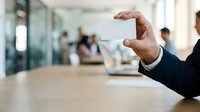 Businessman holding blank white card in modern office corridor with colleagues in background, suitable for corporate branding and business communication concepts.
