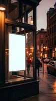 Blank white poster mockup in illuminated storefront window at dusk on urban street with warm lighting and historic brick buildings for advertising display.
