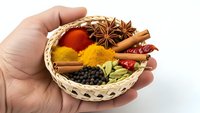 Hand holding wicker basket filled with colorful spices including turmeric, paprika, cinnamon sticks, star anise, peppercorns and cardamom pods.