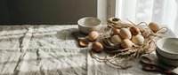 Fresh brown eggs in wicker basket with ceramic bowls on rustic wooden table, natural morning light creating cozy farmhouse kitchen atmosphere for cooking.
