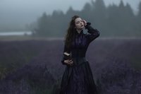 Young woman with long curly hair in dark dress standing in purple lavender field during misty evening with forest background for romantic lifestyle content.