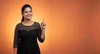 Young Indian businesswoman in elegant black lace dress pointing upward with finger against warm orange gradient background for corporate presentations.
